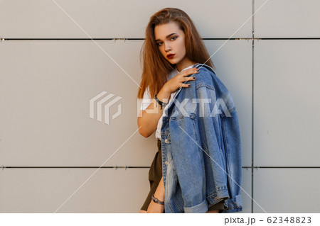Charming stylish young woman in a blue stylish denim jacket in a white  sports dress with a black pattern poses near a white wall. Cute blonde girl  travels. Stock Photo | Adobe